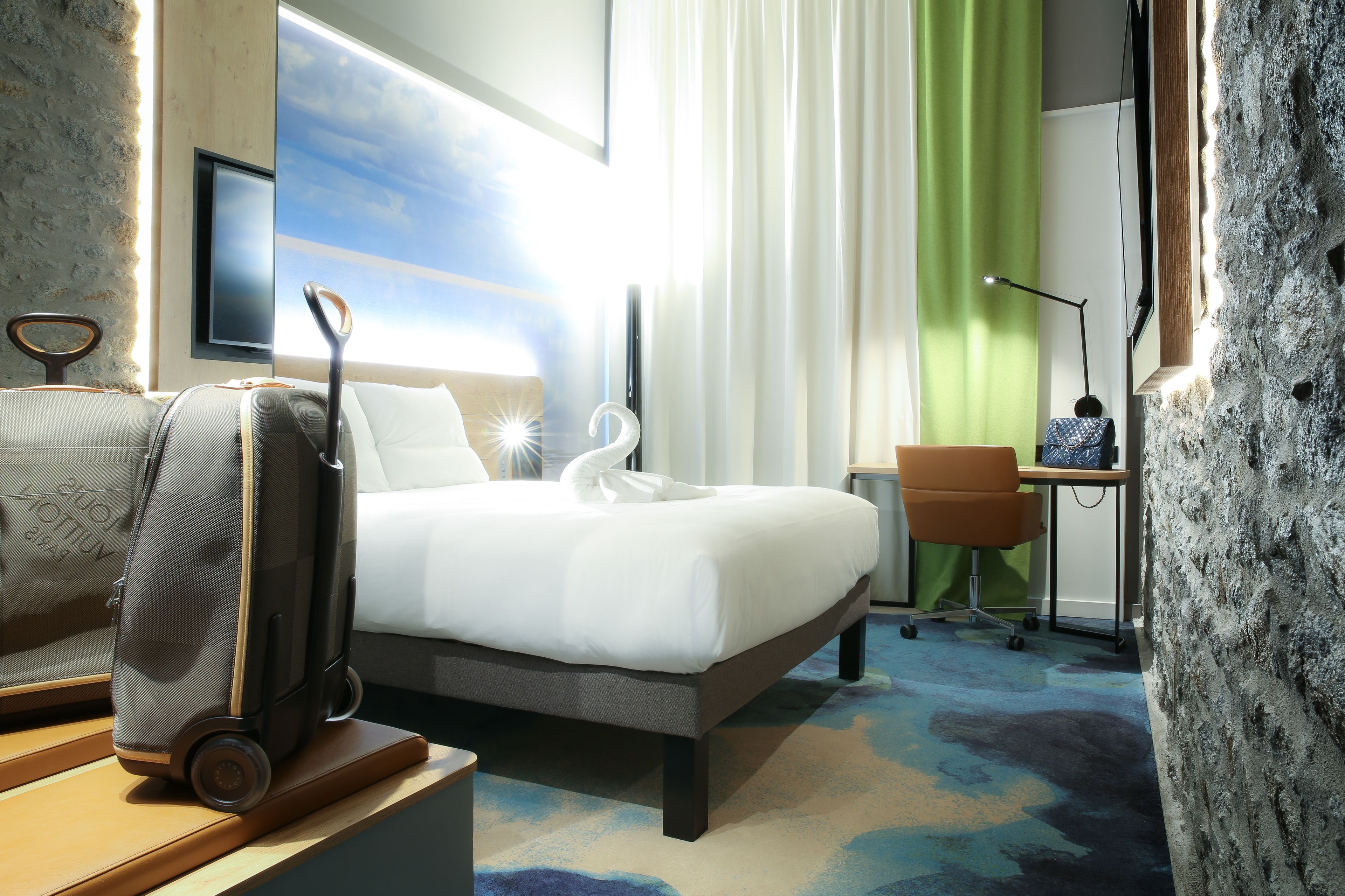 Een hotelkamer met een bed, een bureau, een stoel, een koffer en uitzicht op de lucht.