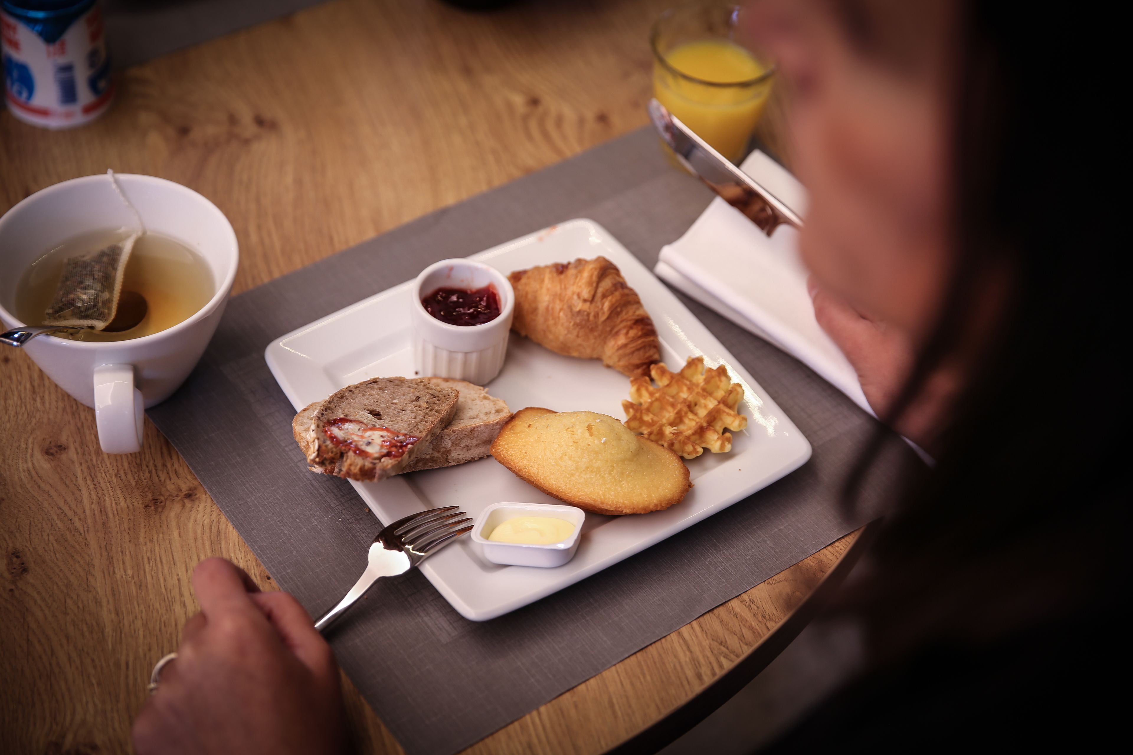 Un plato de desayuno con croissant, mermelada, mantequilla, una taza de té y un vaso de zumo de naranja sobre una mesa de madera.