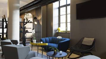 A modern living room with a blue sofa, grey armchairs, and yellow ottomans. There is a round table with a lamp, a TV mounted on the wall, and a window with natural light.