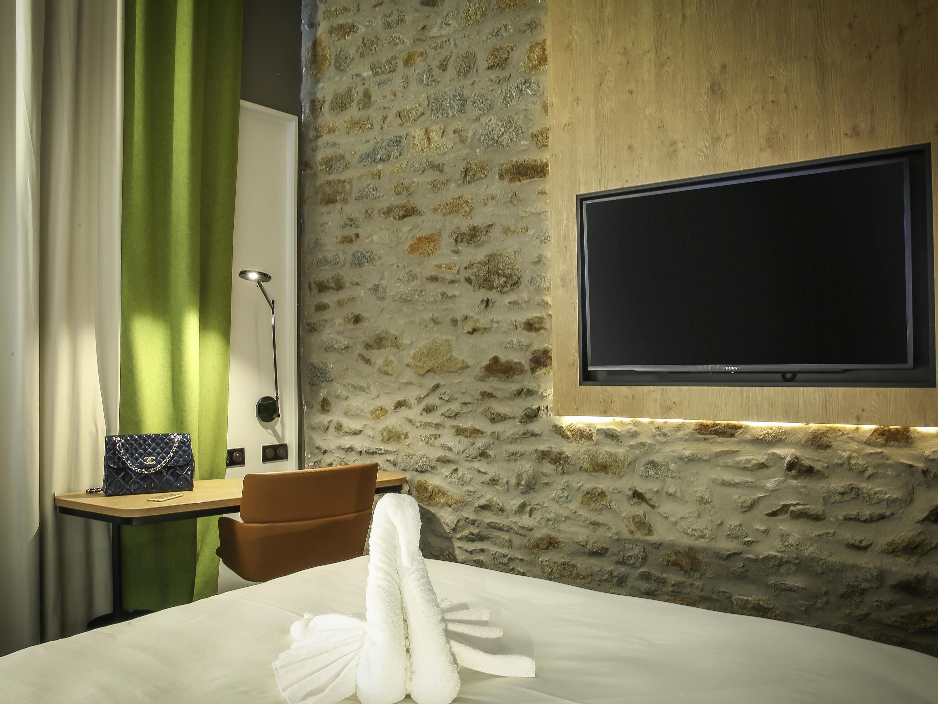 A hotel room with a stone wall, a flat-screen TV mounted on it, a bed with white linens, a green curtain, a small table with a lamp, and a blue handbag.