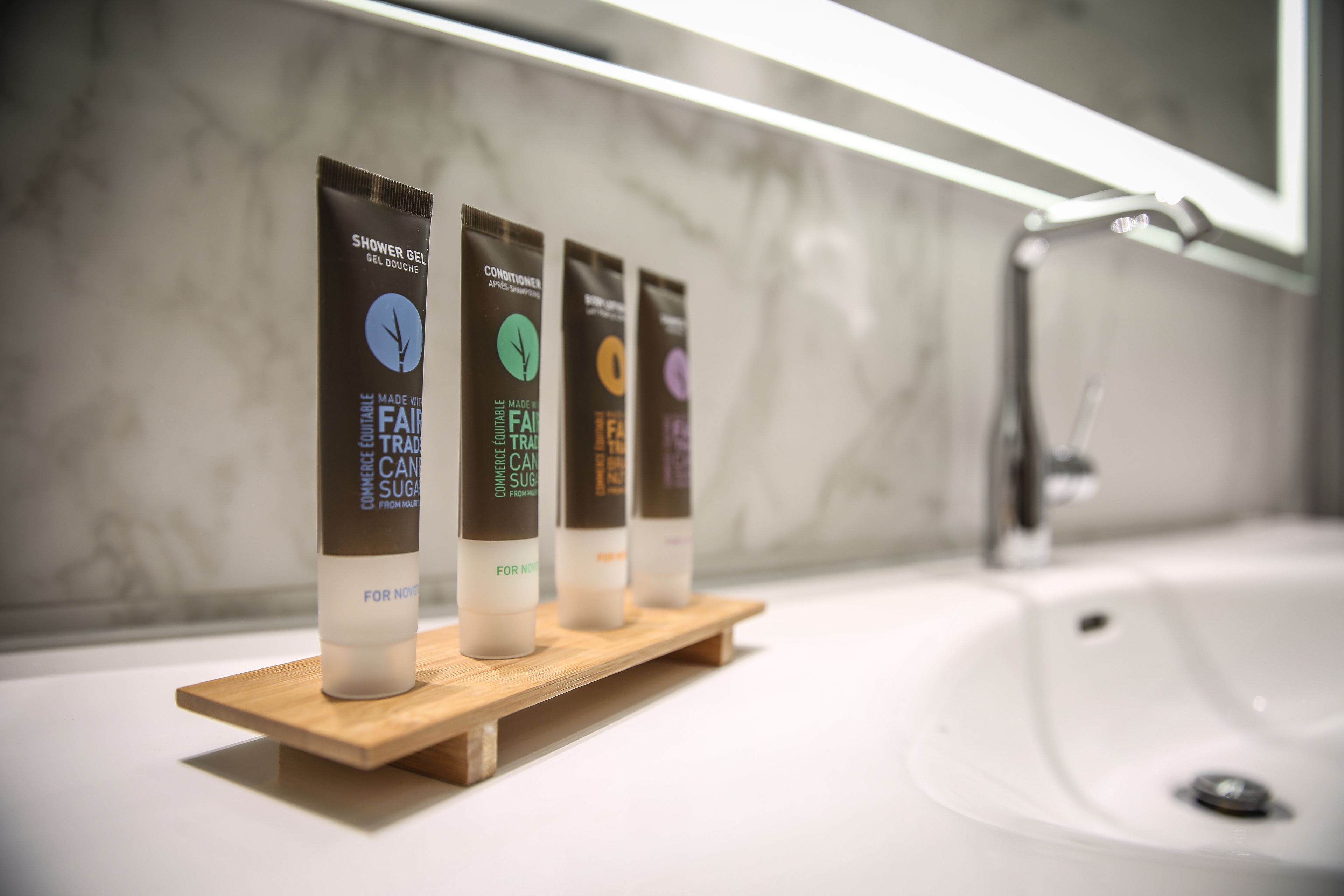 Un baño con pared de mármol, un estante de madera con cuatro botellas de gel de ducha y un lavabo blanco con grifo.