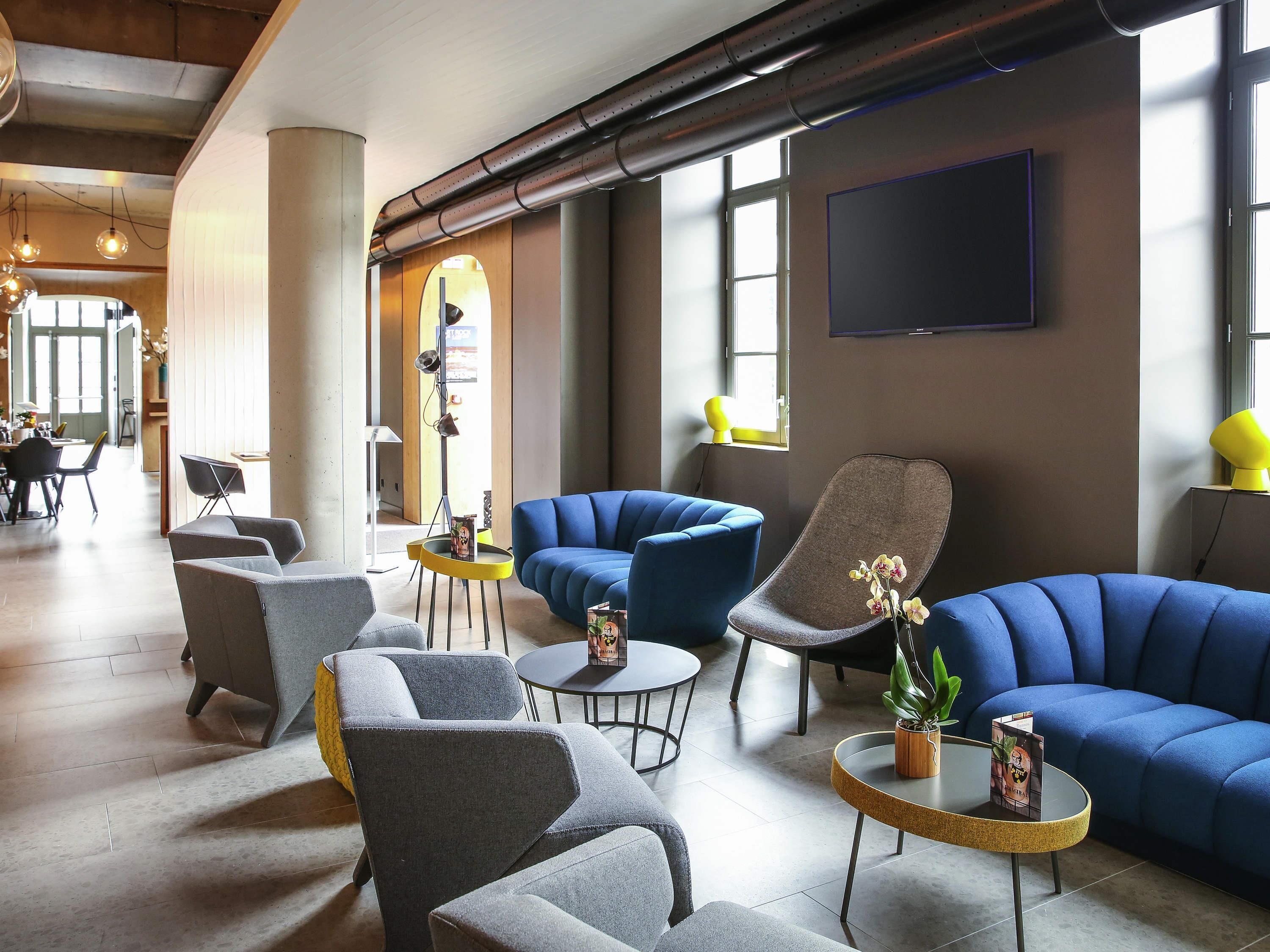 A modern lounge area with sofas, armchairs, and tables, featuring a large window and a flat-screen TV.