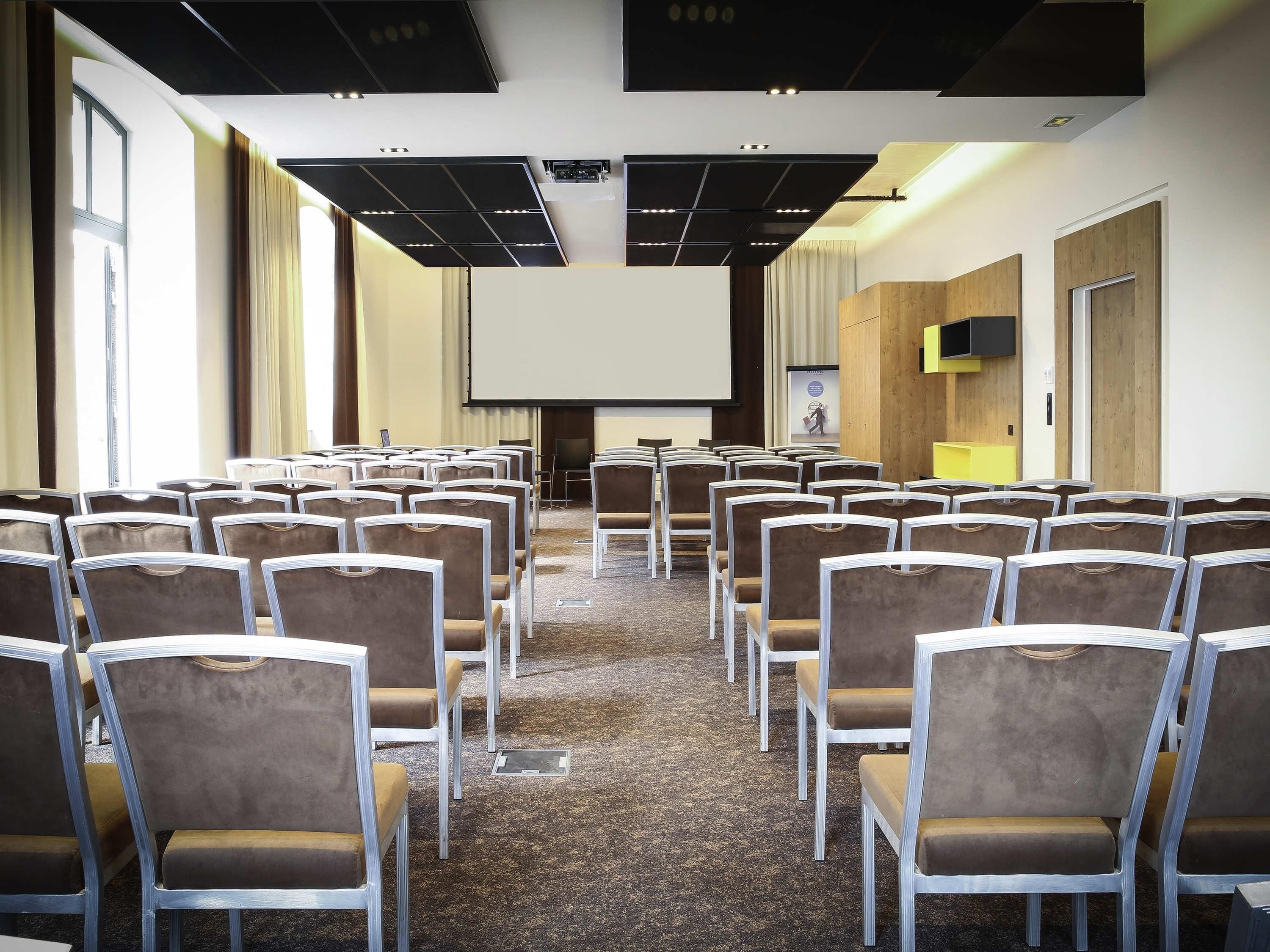 A conference room with rows of chairs, a projector screen, and a flat-screen TV on the wall.