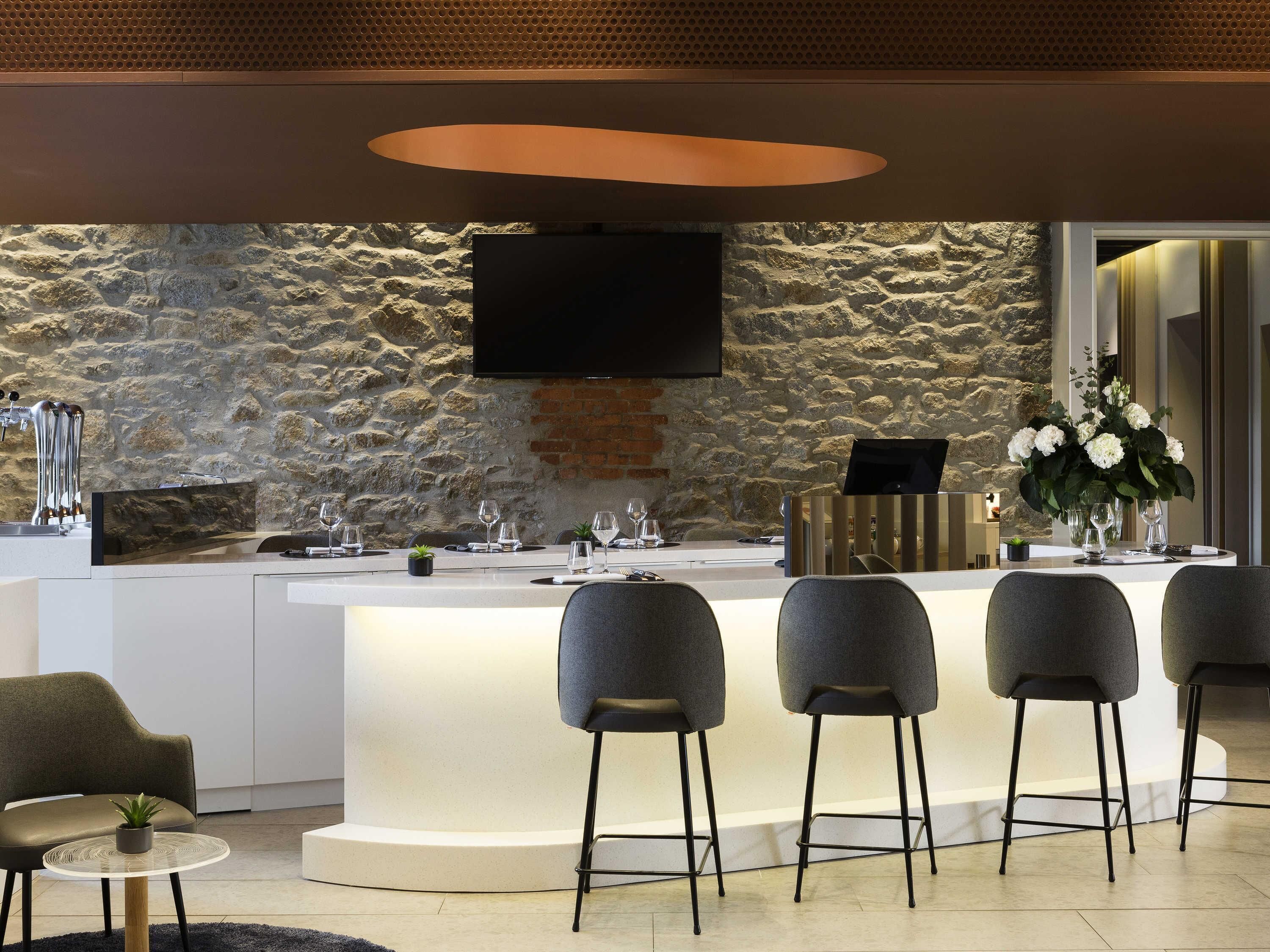 Een moderne barruimte met een achtergrond van een stenen muur, een flatscreen televisie boven de bar en een zithoek met barstoelen.