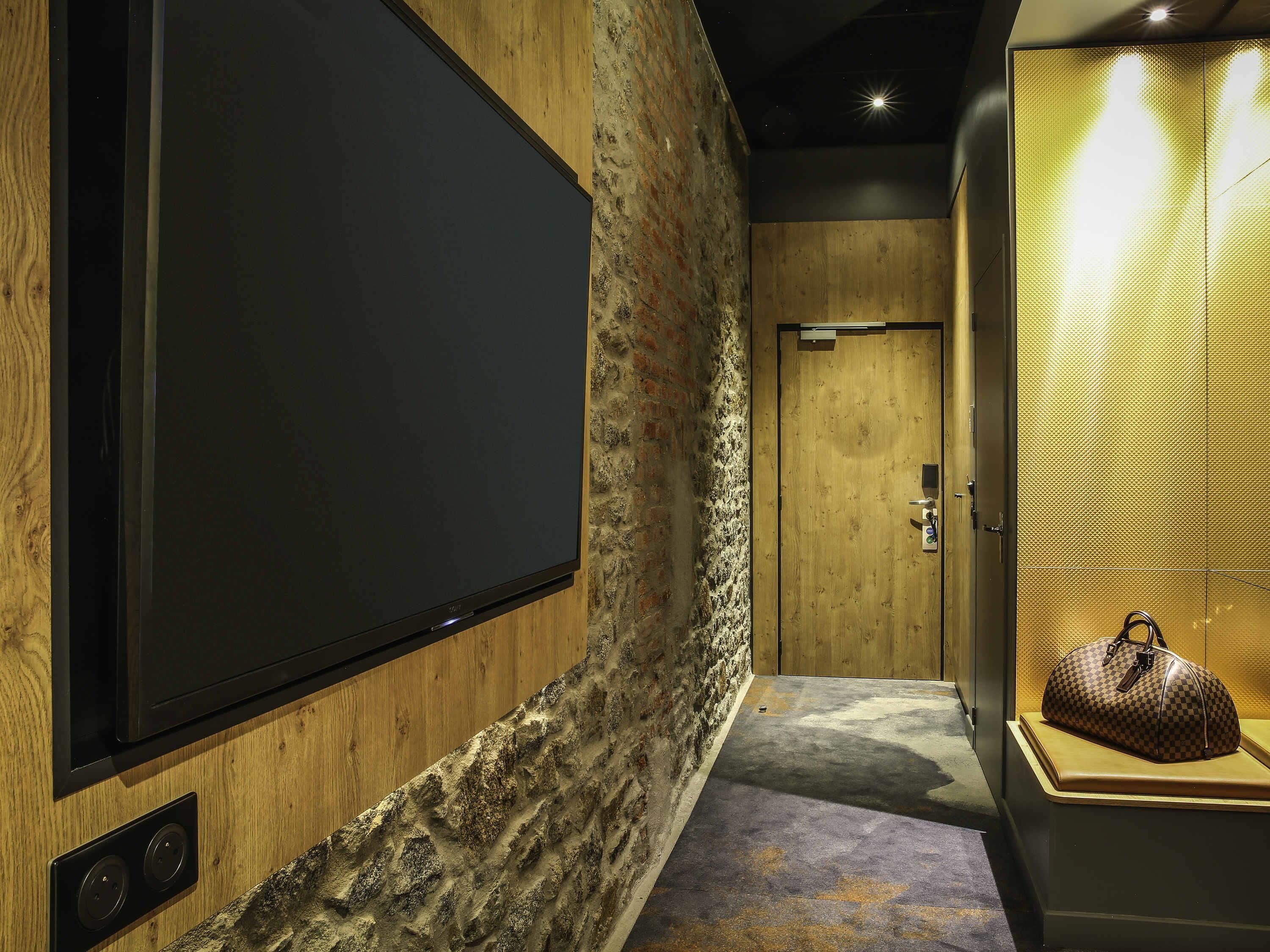 A hotel room with a large flat-screen TV mounted on a wooden wall, a stone-textured wall, a wooden door, and a small display case with a handbag.
