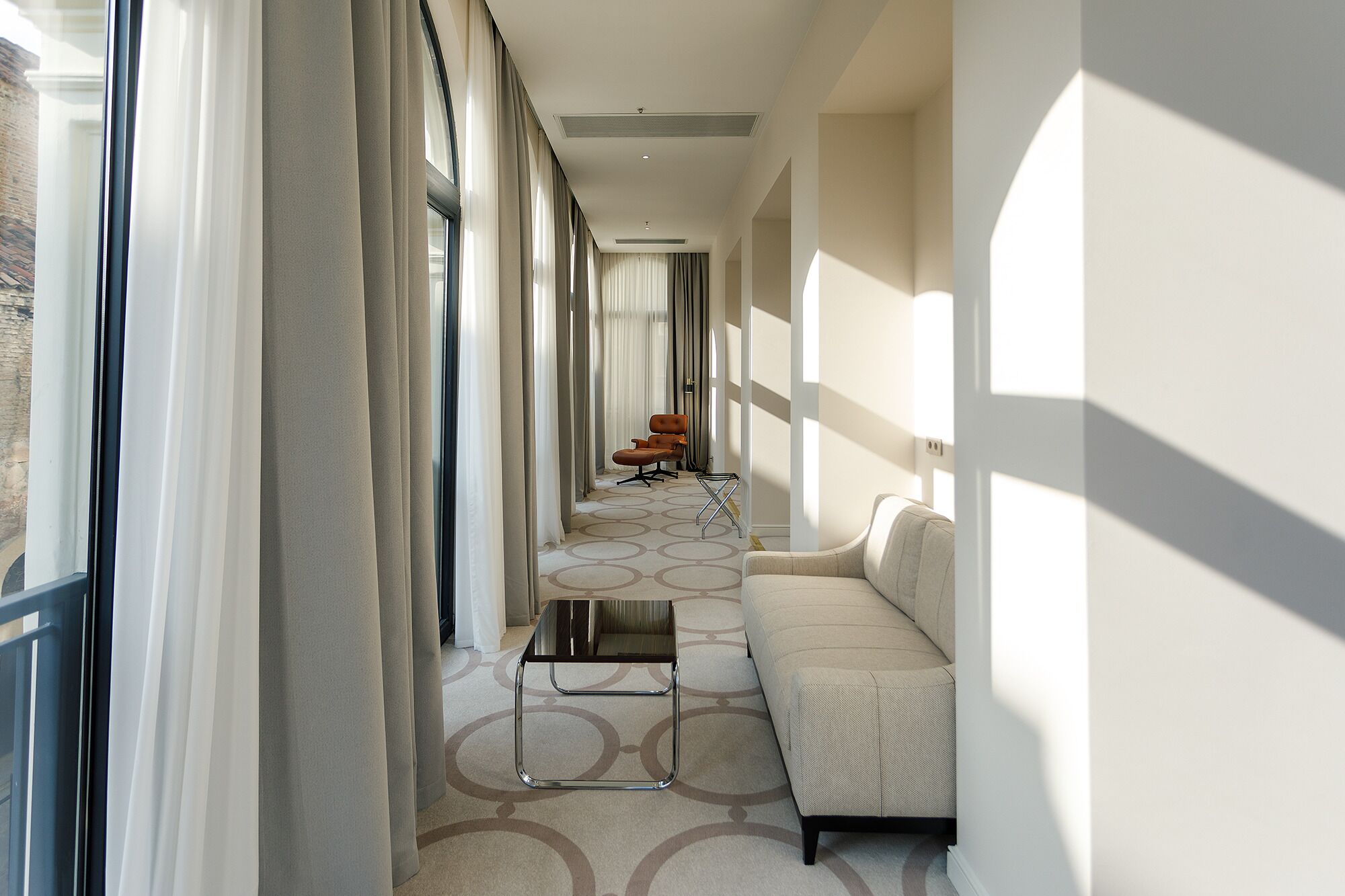 A corridor with a sofa, a chair, and a table, leading to a window with curtains.