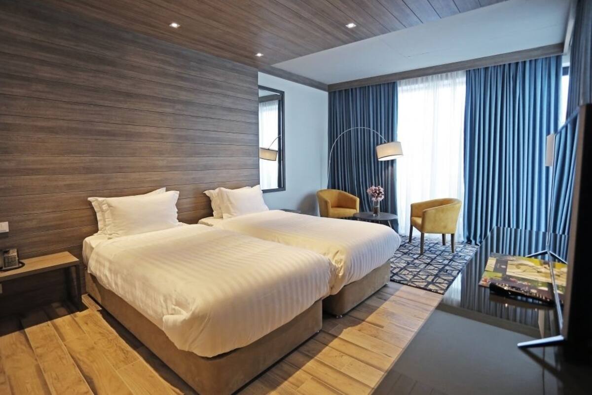 Habitació d'hotel amb dos llits, un escriptori amb una làmpada, una cadira i un finestral gran amb cortines.