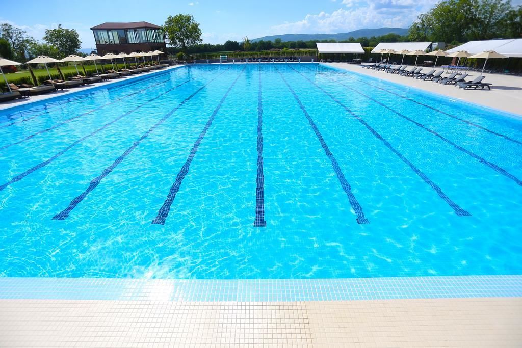 Ein großer, klarer, blauer Schwimmteich mit mehreren Bahnen, umgeben von Liegestühlen und Sonnenschirmen, mit einem Gebäude im Hintergrund.