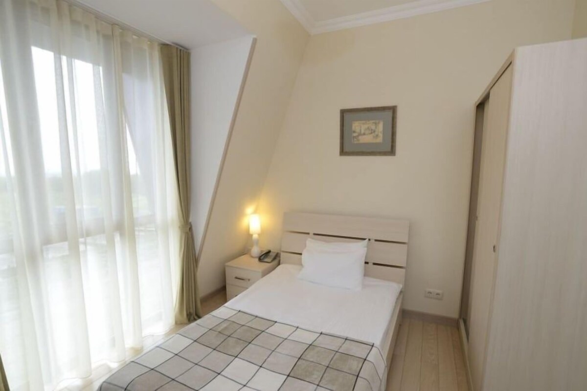 A bedroom with a bed, a nightstand with a lamp, a framed picture on the wall, and a window with curtains.