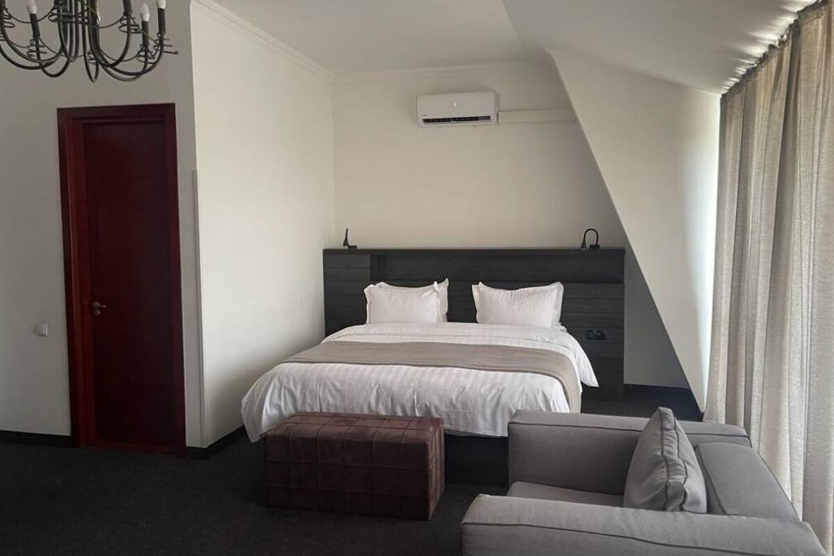 Una habitació d'hotel moderna amb un llit gran, un panell de capçal fosc, una porta de fusta, un otomà petit, un sofà gris i una làmpada penjant.
