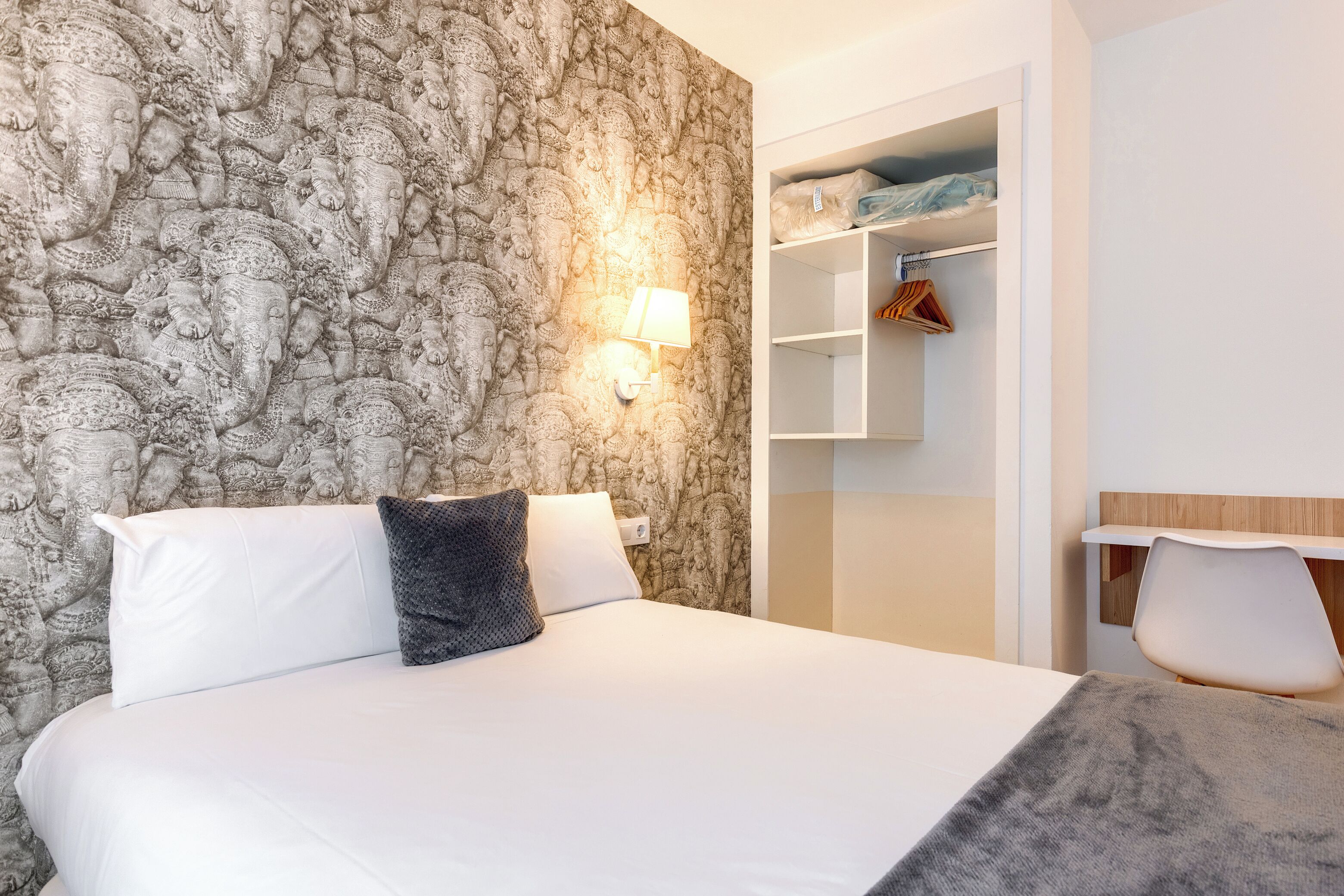 A hotel room with a large bed, a nightstand, a wall-mounted lamp, and a wardrobe with hanging clothes.