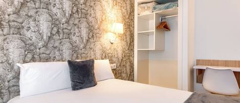 A hotel room with a large bed, a nightstand, a wall-mounted lamp, and a wardrobe with hanging clothes.