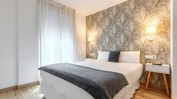 Quarto de hotel com uma cama grande, duas luminárias de cabeceira, parede com textura de pedra, piso de madeira e uma janela com cortinas.