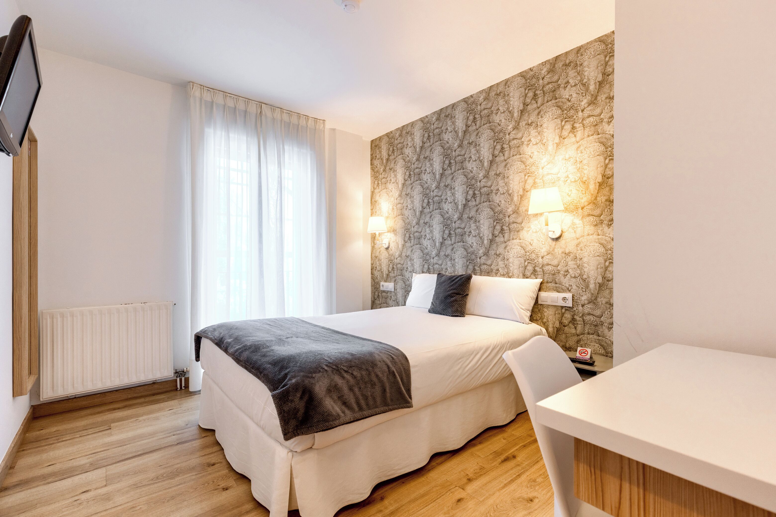 Quarto de hotel moderno com cama, escrivaninha, cadeira, janela com cortinas, parede de pedra e luminária na parede.