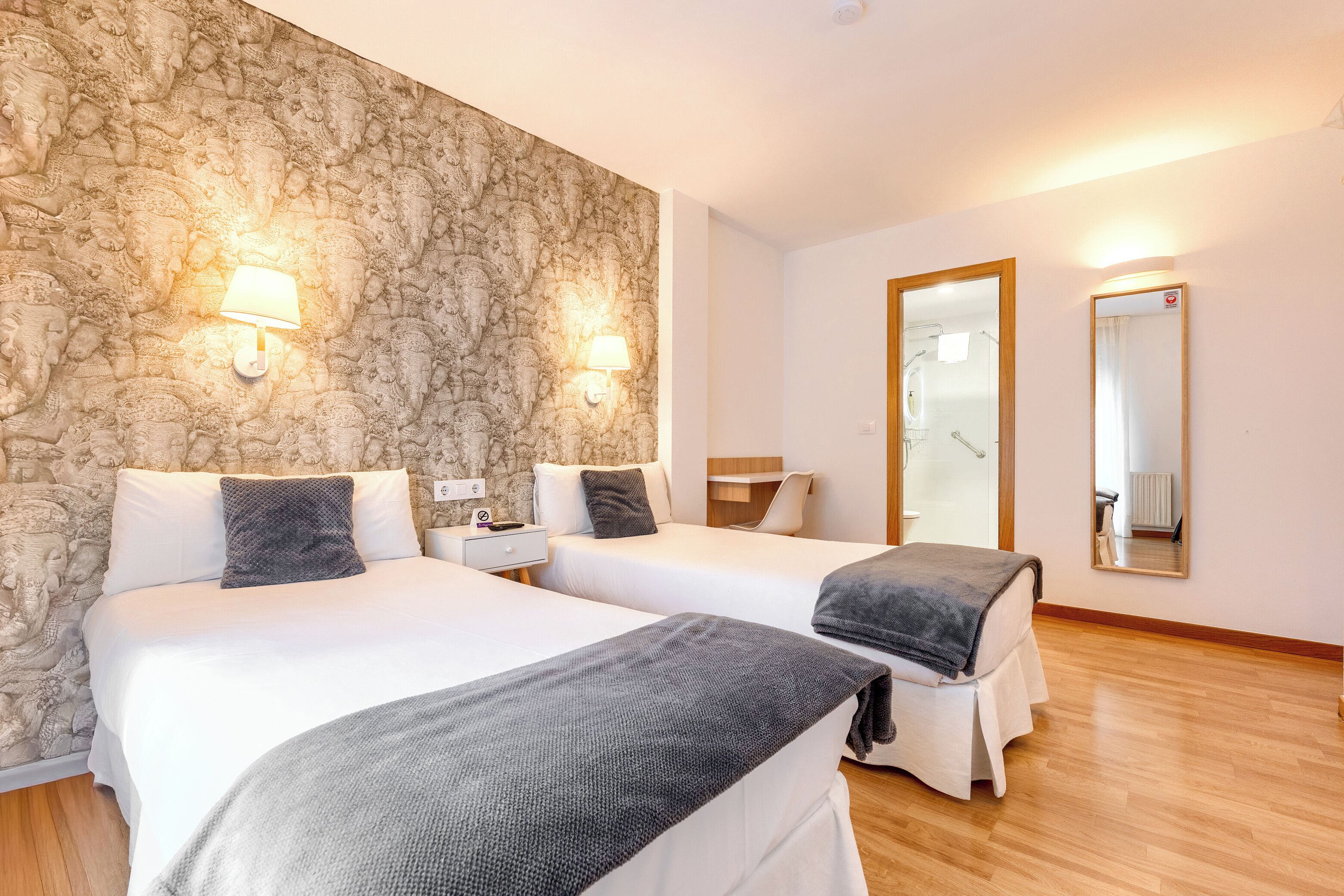 Une chambre d’hôtel avec deux lits, un mur décoré d’un motif en pierre, un plancher en bois, un miroir et une table de chevet avec une lampe.