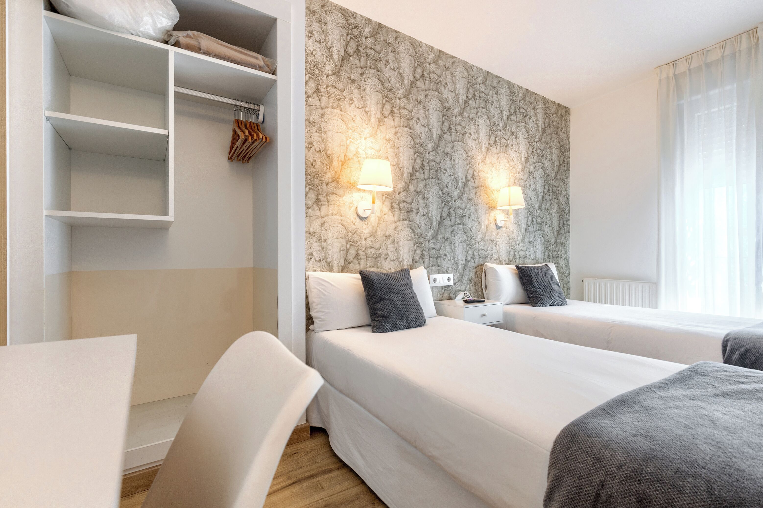 A hotel room with two beds, a wardrobe, a desk, and a wall with a textured pattern.