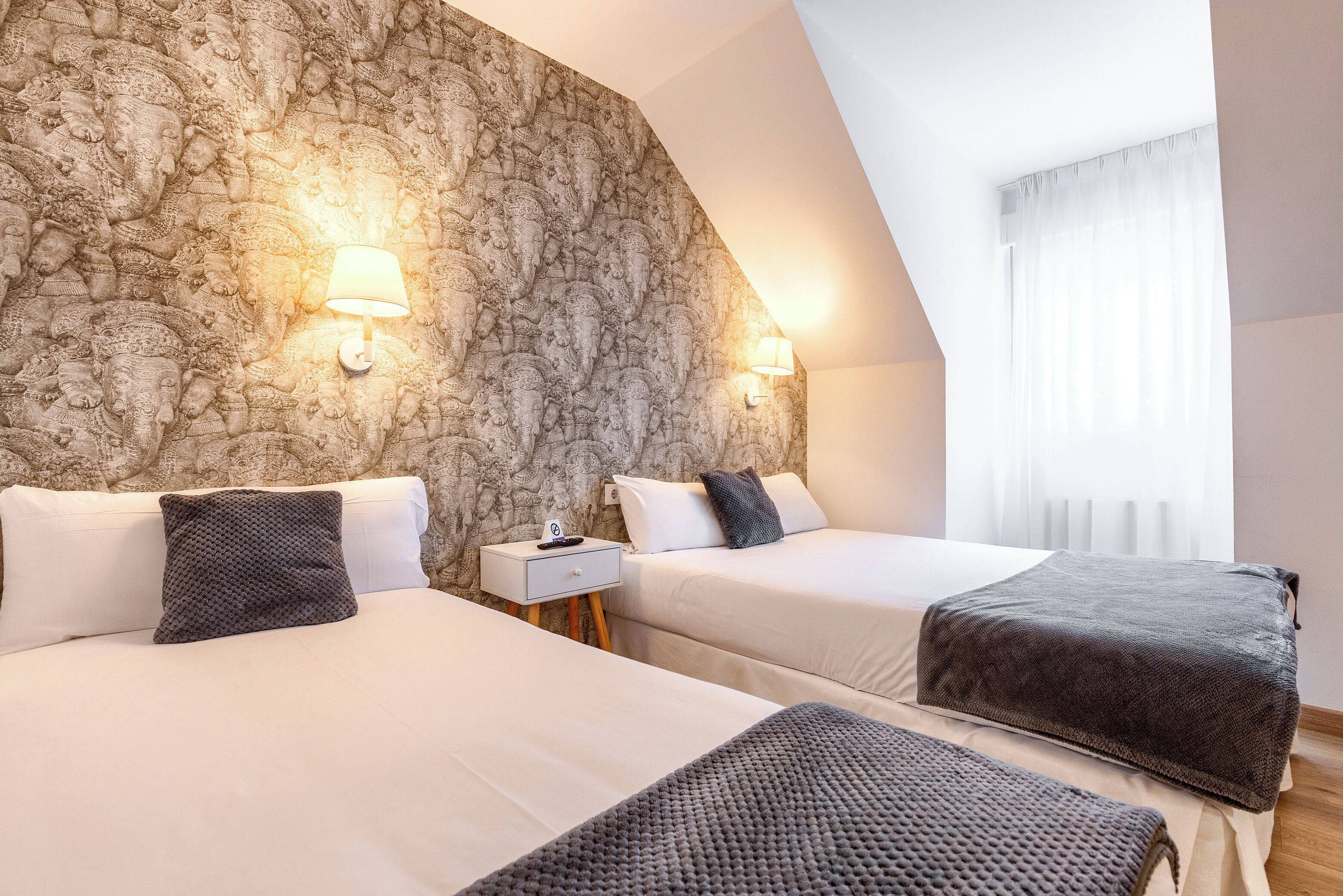 A hotel room with two beds, a stone wall, two wall-mounted lamps, a nightstand, and a window with curtains.