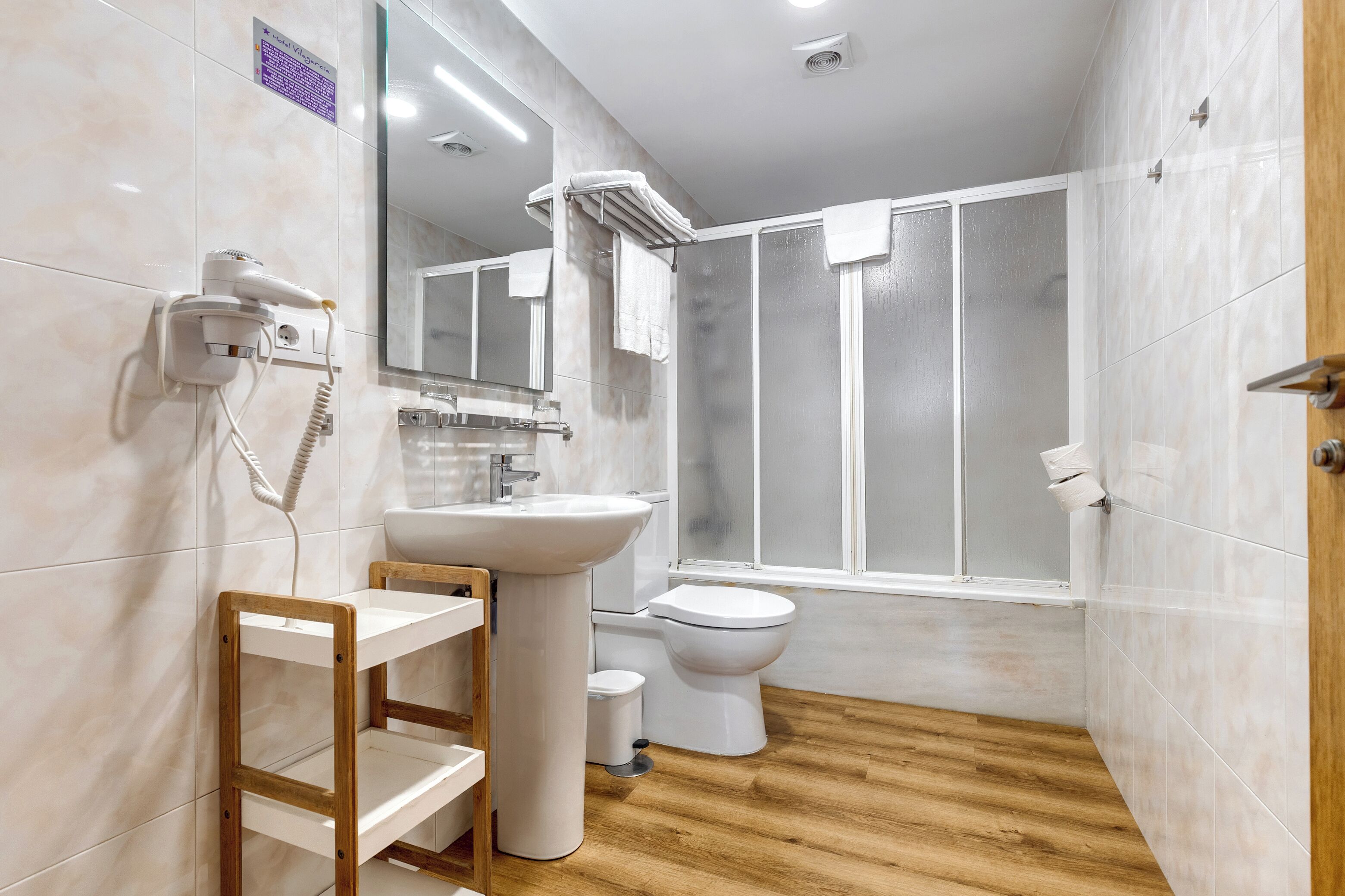Une salle de bain avec un plancher en bois, des carreaux blancs, un lavabo, des toilettes et une douche dotée de portes en verre dépoli.