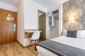Habitación de hotel con puerta de madera, un escritorio y una silla, cama con manta, armario y lámpara fijada a la pared.
