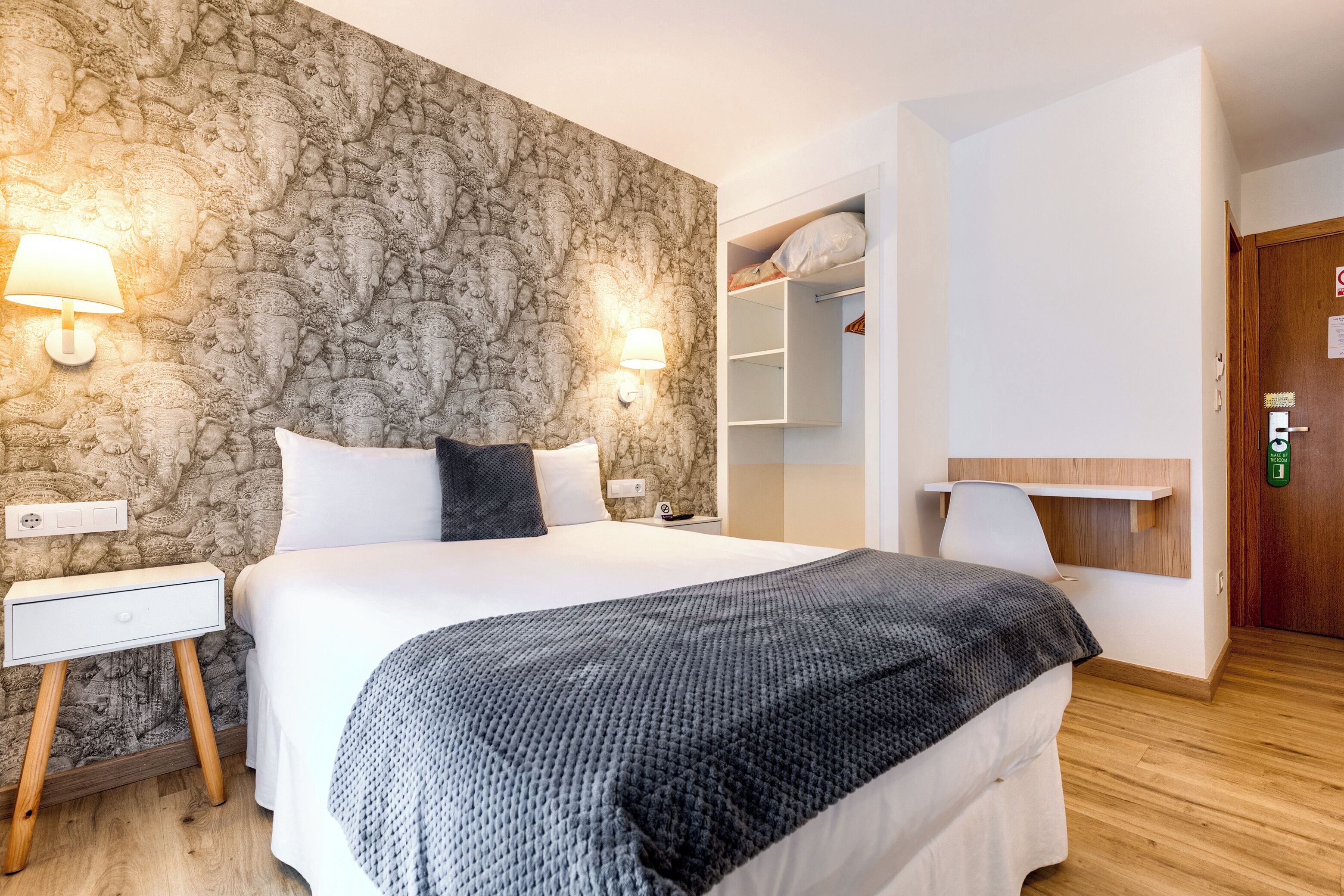 Quarto com parede em padrão de pedra, cama com roupa de cama branca, mesa de cabeceira, luminária, guarda-roupa e porta com placa de saída de emergência verde.