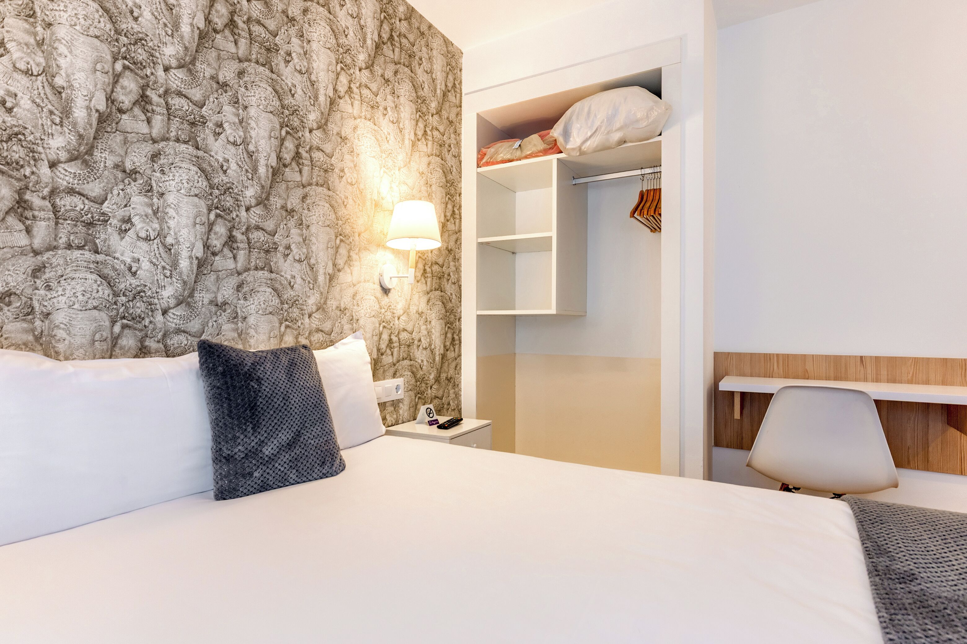 A hotel room with a bed, a nightstand, a lamp, a wardrobe, and a chair.