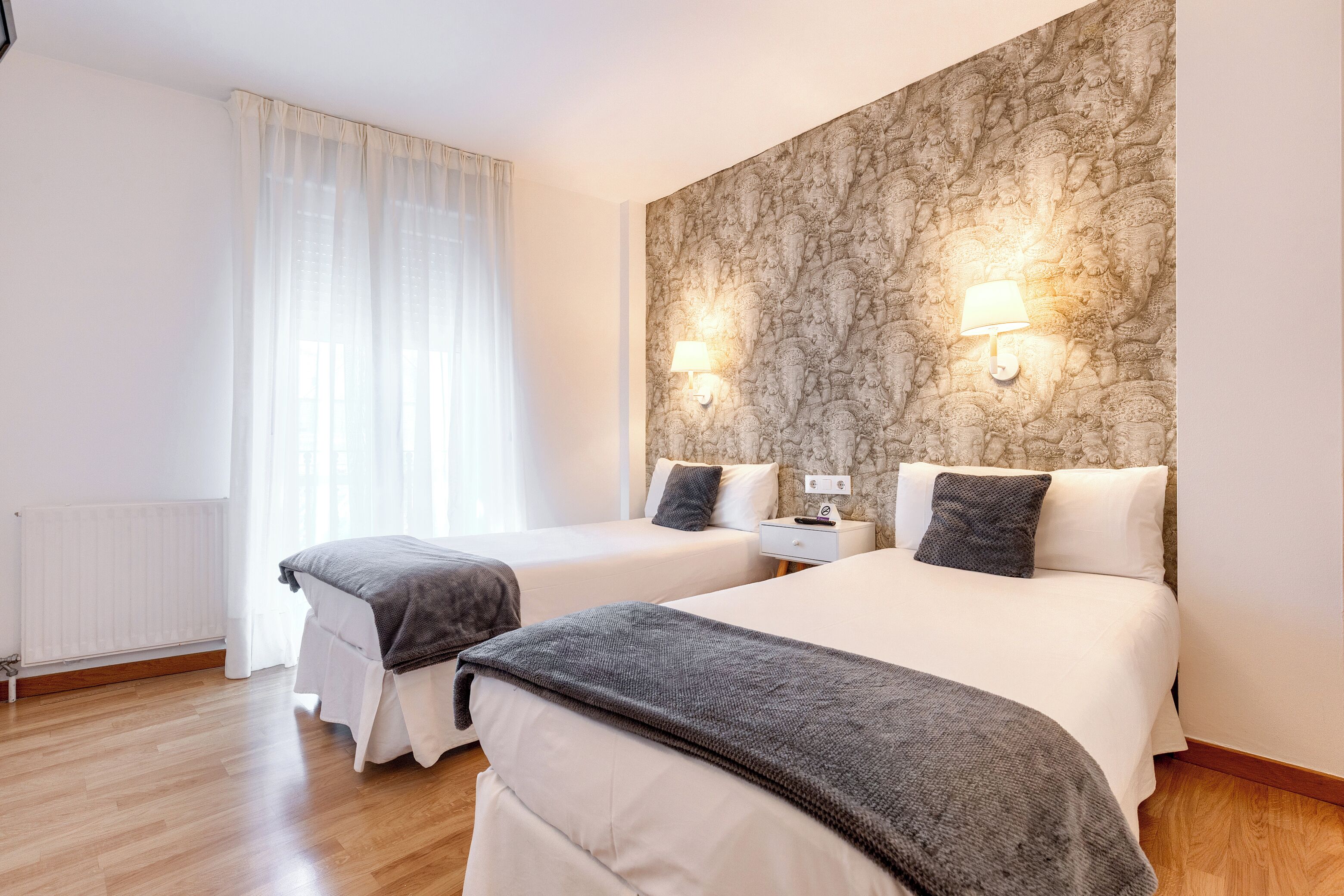 Ein Hotelzimmer mit zwei Betten, einem Heizkörper, einem Fenster mit durchsichtigen Vorhängen und einer strukturierten Wand.