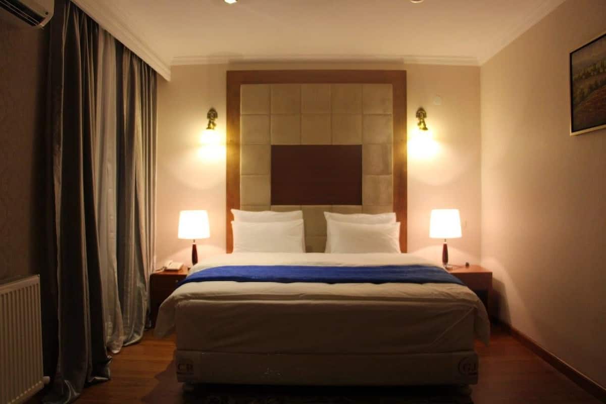 Una habitación de hotel con una cama grande, dos lámparas de noche, un cabecero y una ventana con cortinas.