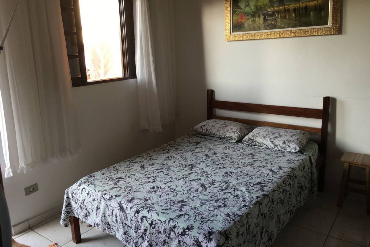 Une chambre à coucher avec un lit, une fenêtre avec des rideaux, un tableau accroché au mur et un petit tabouret en bois.