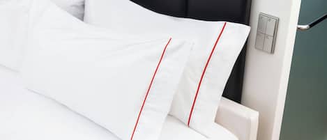 Una cama bien hecha con ropa blanca y ribetes rojos, un cabecero negro y un interruptor de luz en la pared.