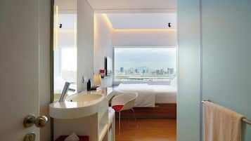 A modern hotel room with a large window, a bed, a white chair, and a bathroom with a sink and mirror.