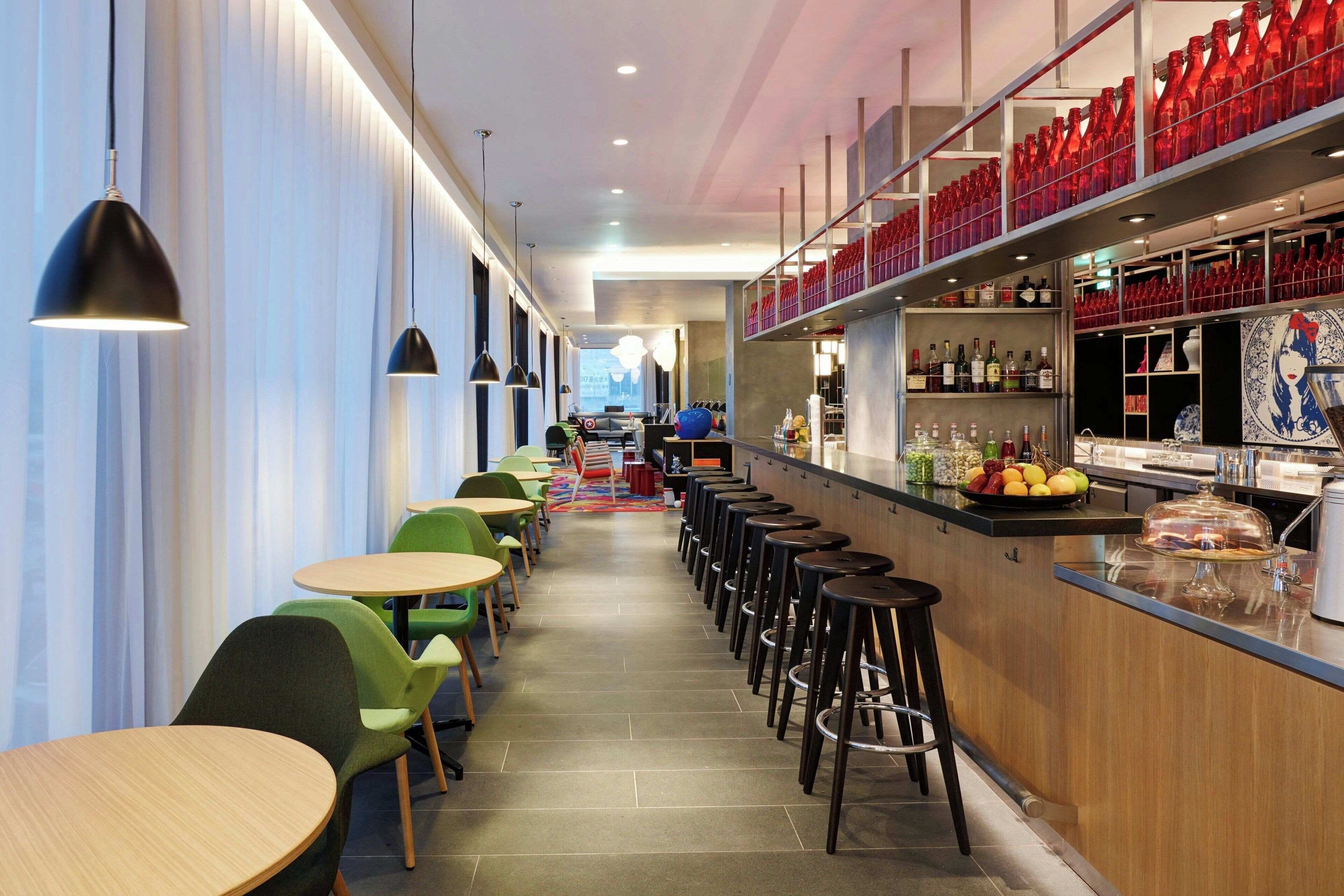 A modern bar area with high stools, a counter with a variety of bottles, and a seating area with round tables and chairs.