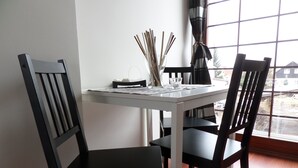 Una mesa de comedor con sillas, un ventanal con vistas a casas y un florero con ramas.