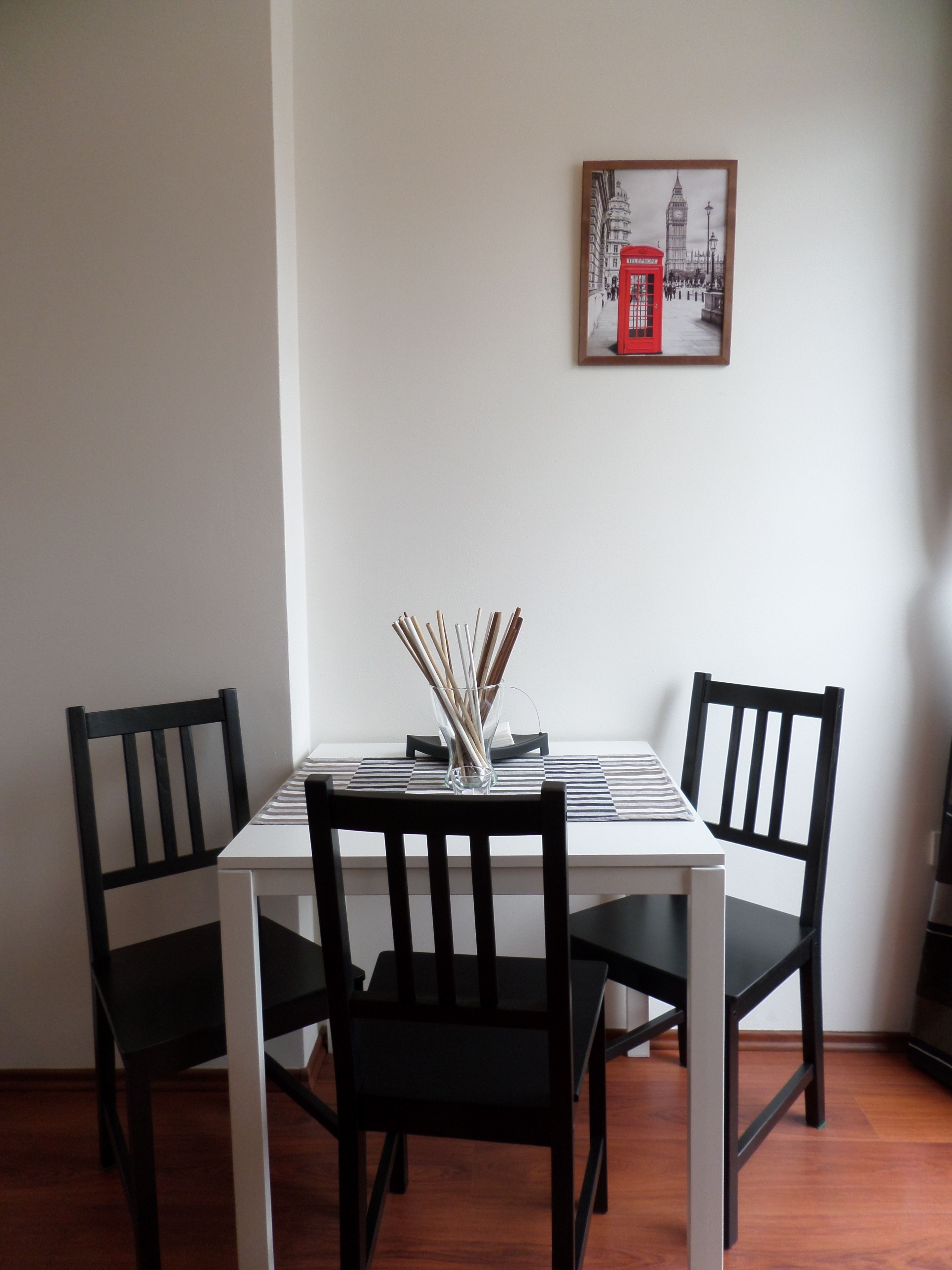 A dining table with four black chairs, a clear glass vase with sticks, and a framed picture of a red telephone booth on the wall.