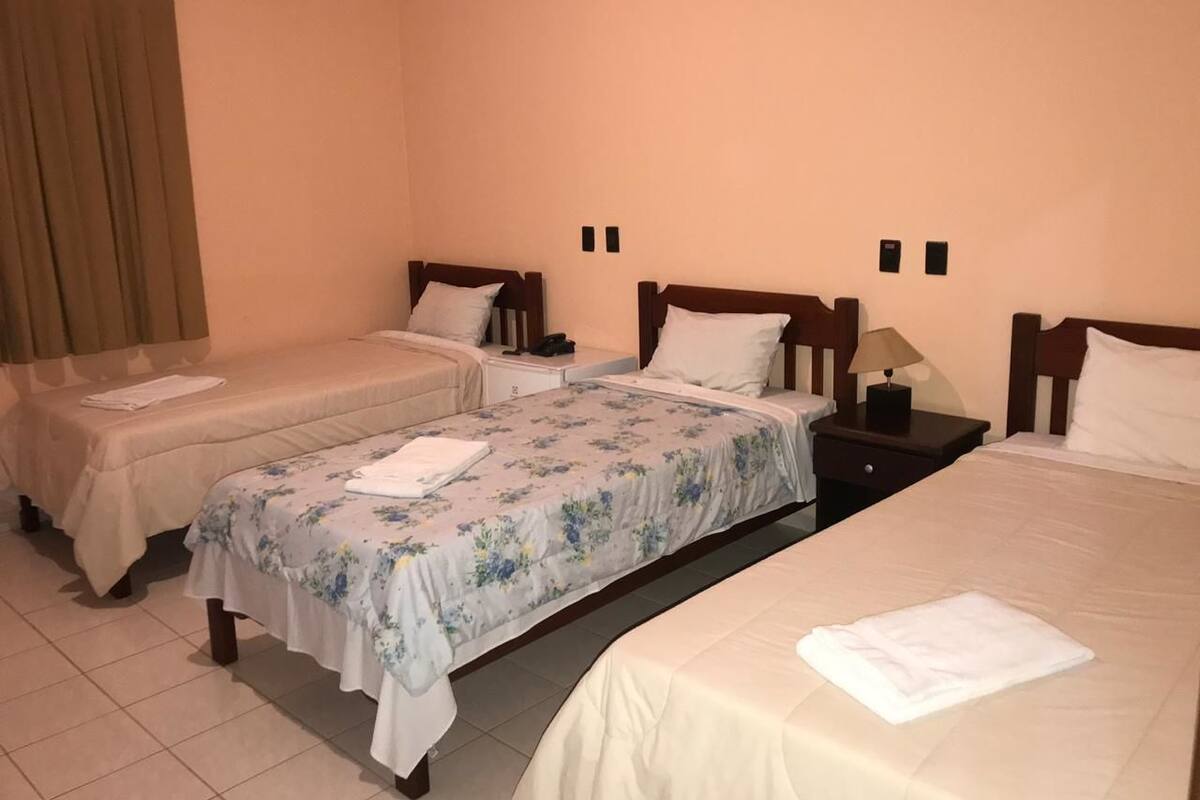 Habitación de hotel con dos camas individuales, una cama más grande y una mesita de noche con lámpara.