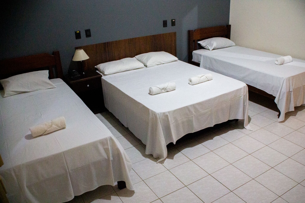 Habitación de hotel con tres camas individuales, cada una con una colcha blanca y toallas enrolladas.