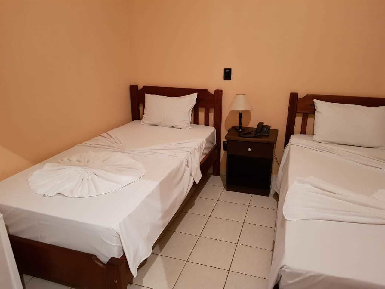 Une chambre avec deux lits simples, chacun doté d’une tête de lit en bois, de draps blancs et d’une petite table de chevet avec une lampe.