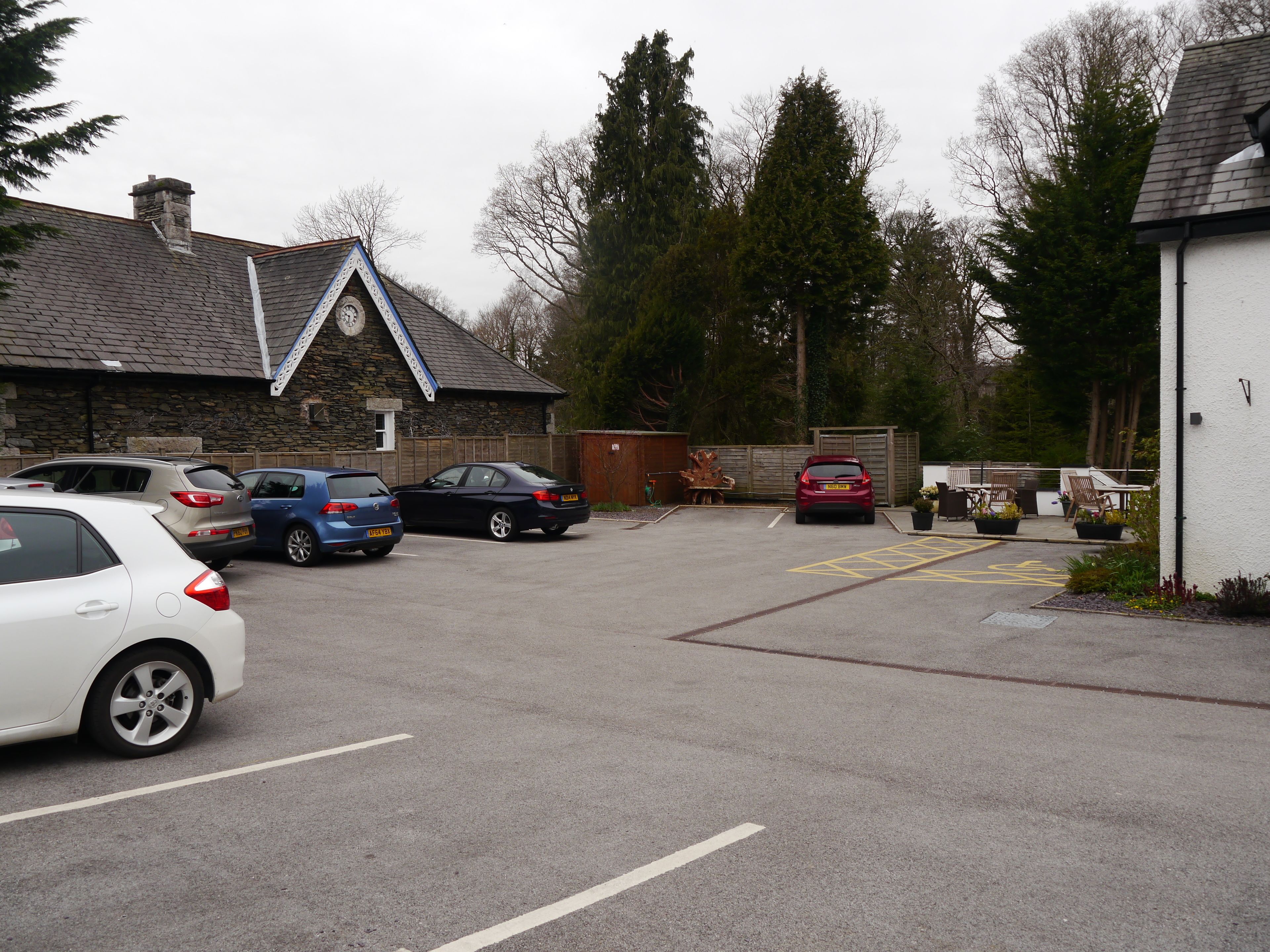A parking area with several cars, a stone building with a triangular roof, and a fenced garden area with benches.