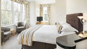 A hotel room with a bed, a TV, a chair, a desk, and large windows.
