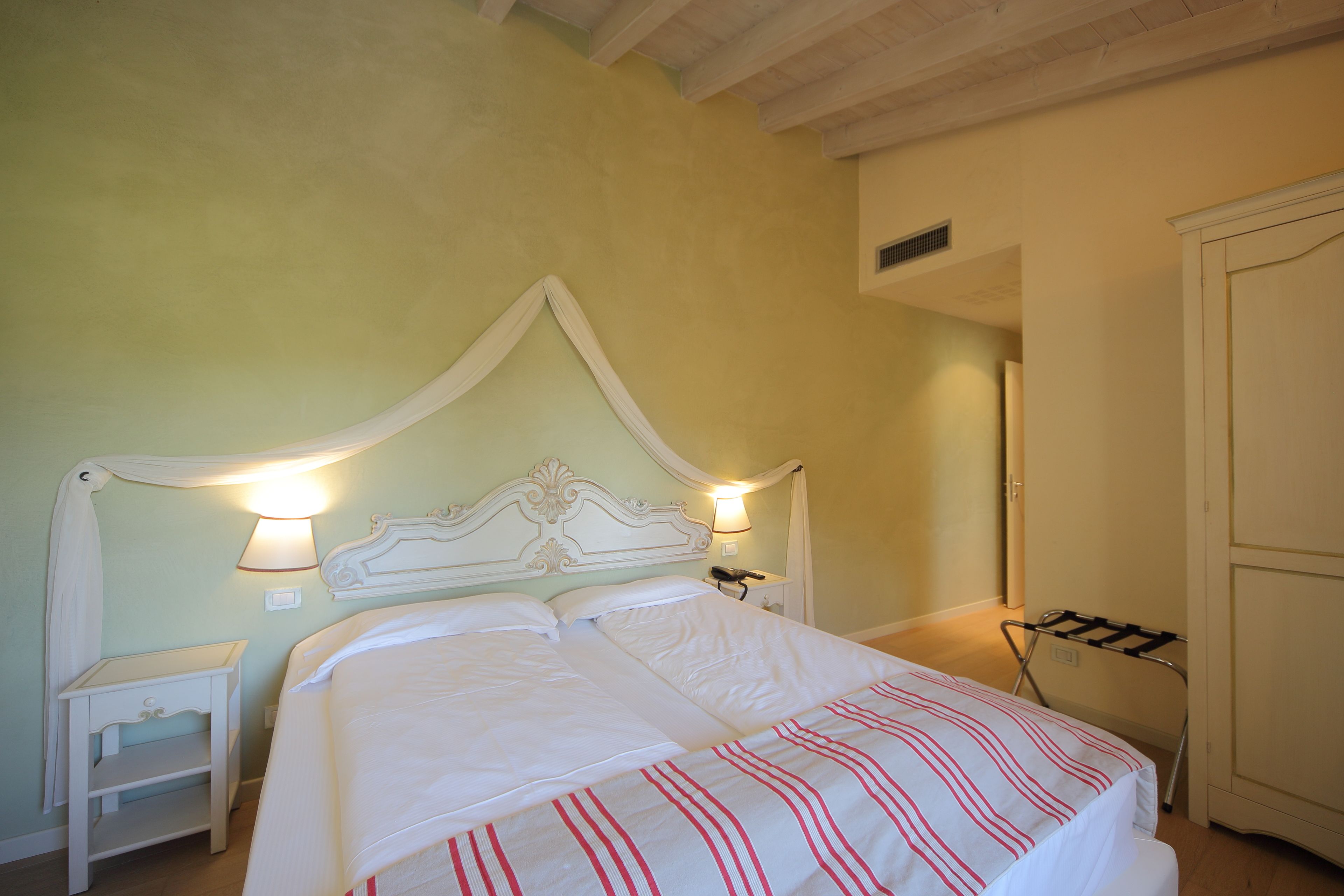 A neatly made bed with white linens and red stripes, a bedside table with a lamp, and a wardrobe in a room with light green walls.
