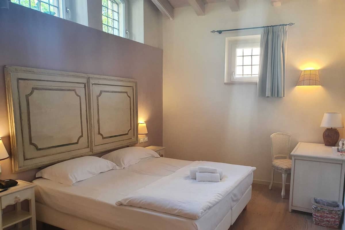 A bedroom with a large bed, two bedside tables, a chair, a small table with a lamp, and a window with curtains.