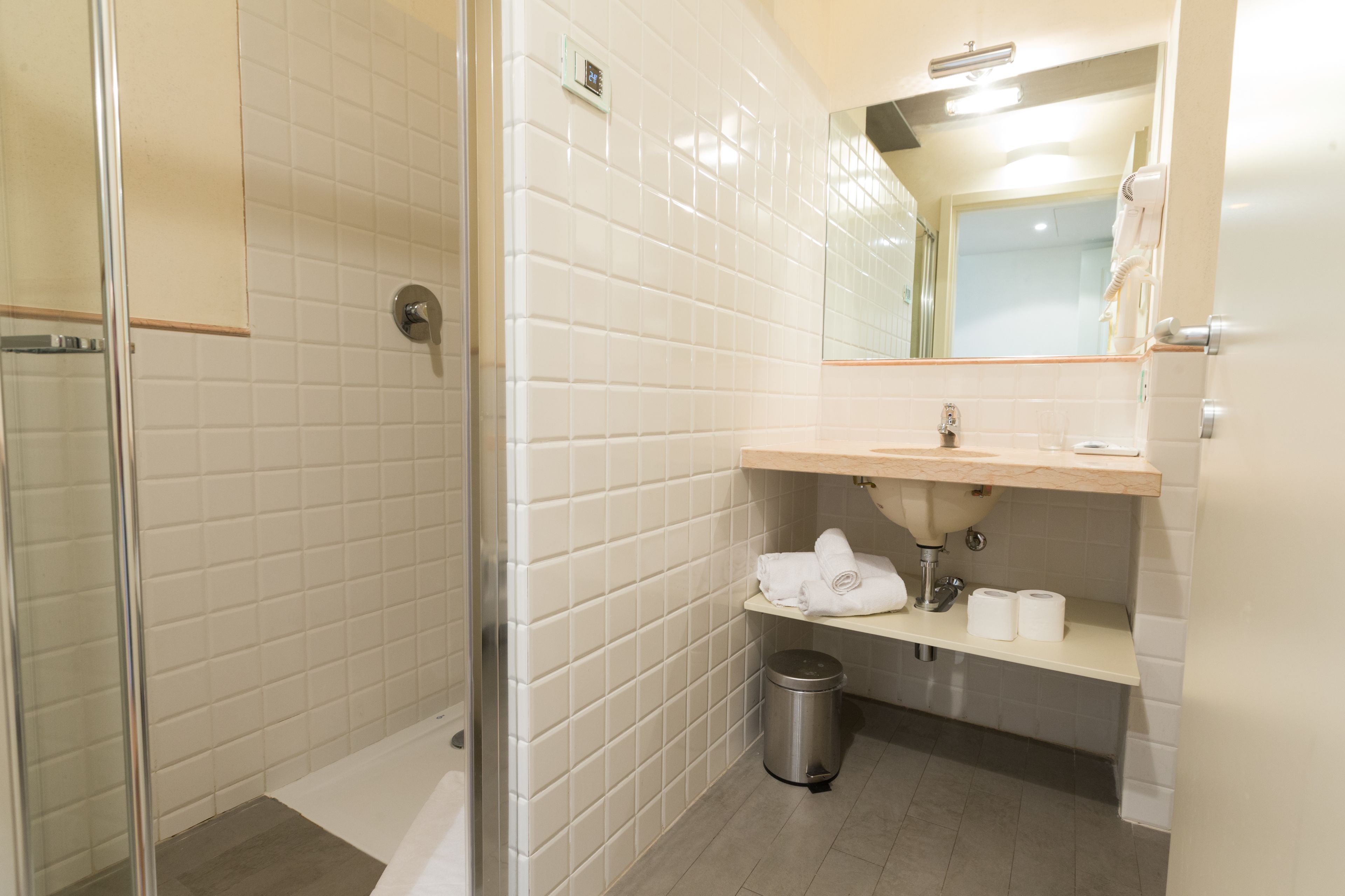 A bathroom with white tiled walls, a glass shower enclosure, a sink with a mirror above it, and a trash can.