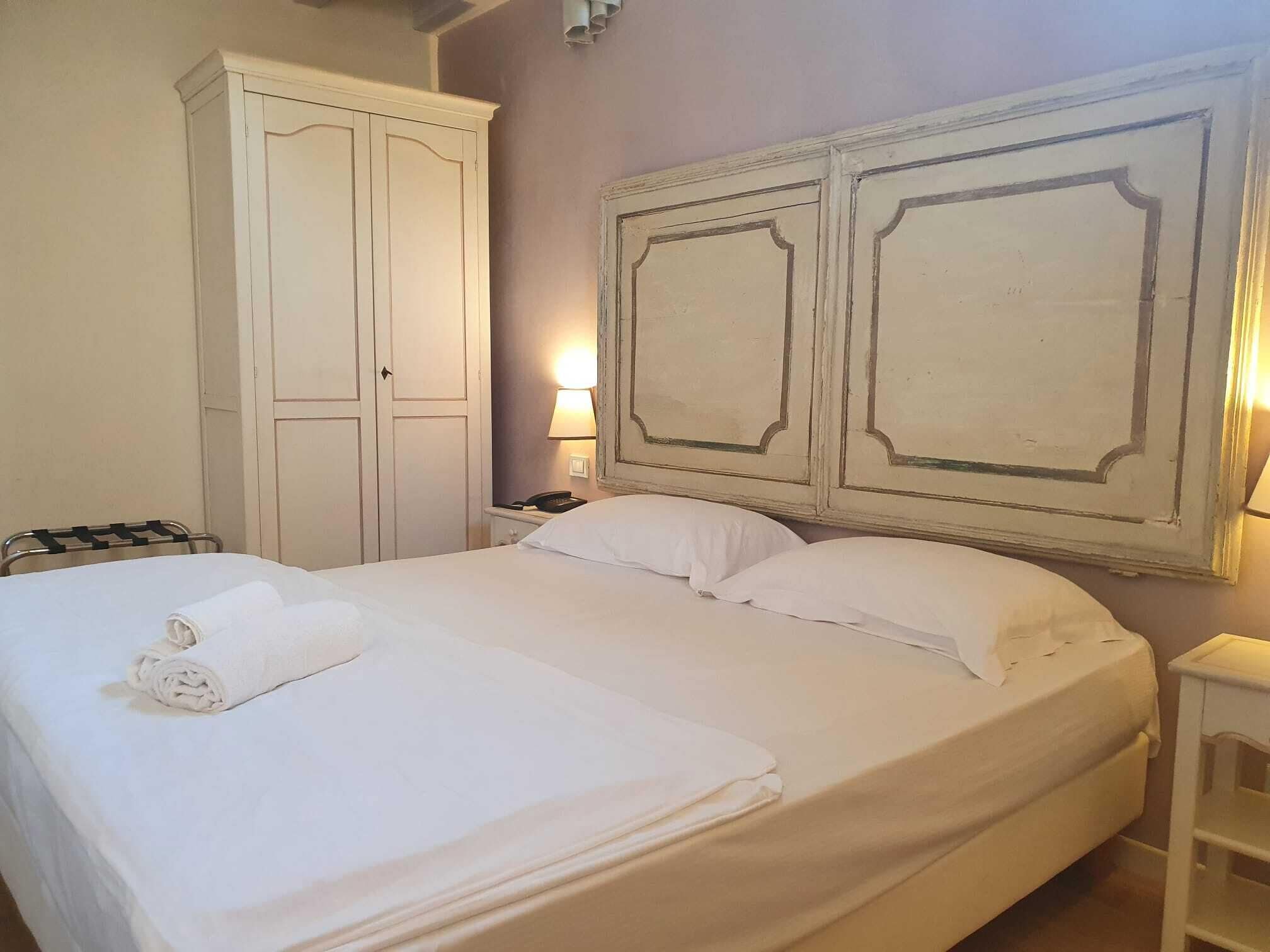 Un lit bien fait, avec du linge de lit blanc et une serviette pliée, une armoire blanche et une table de chevet avec une lampe.