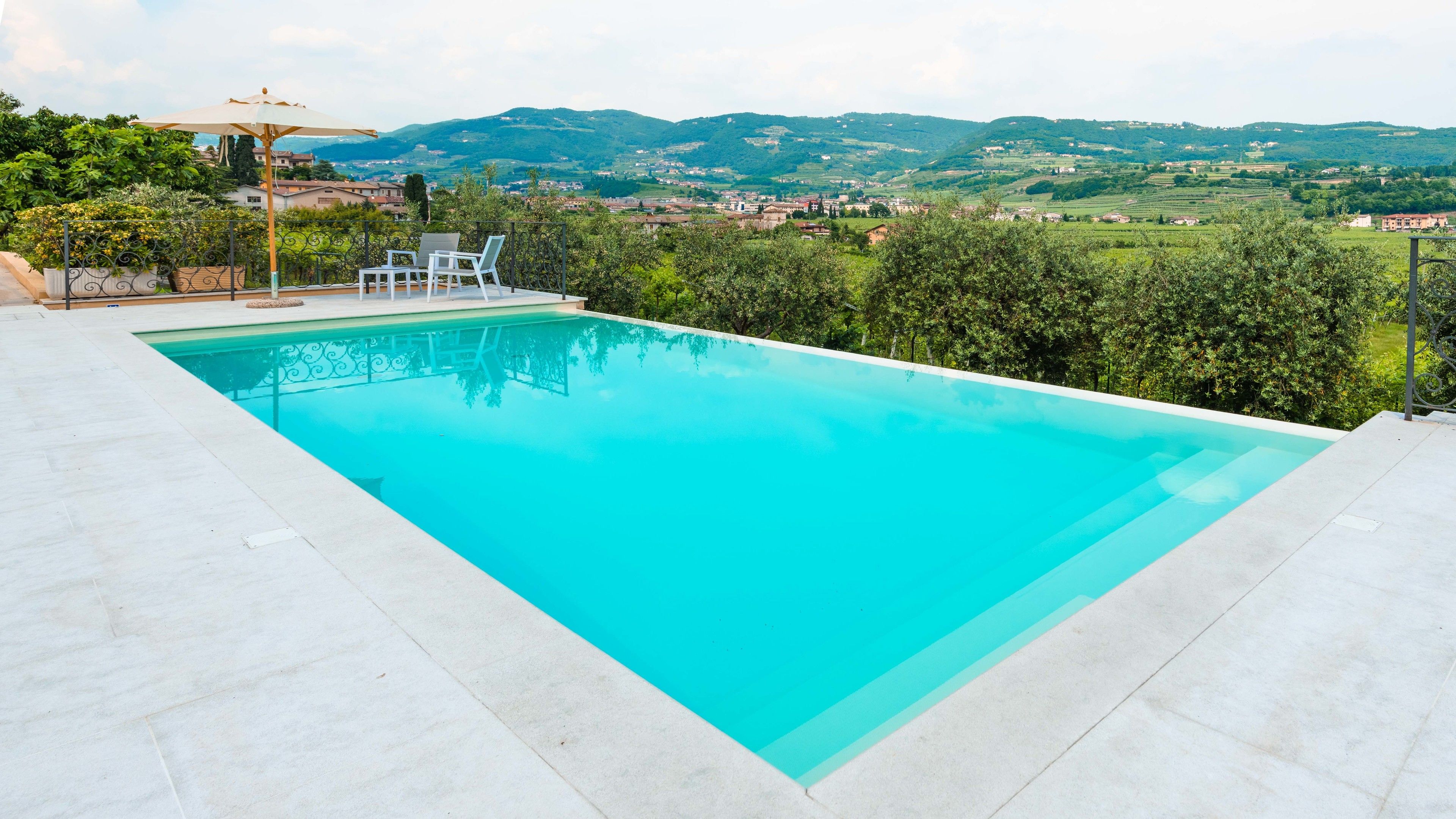 Una piscina rectangular con agua cristalina azul, rodeada por una terraza con baldosas de color claro. Hay dos sillas blancas y un paraguas beige en la terraza.