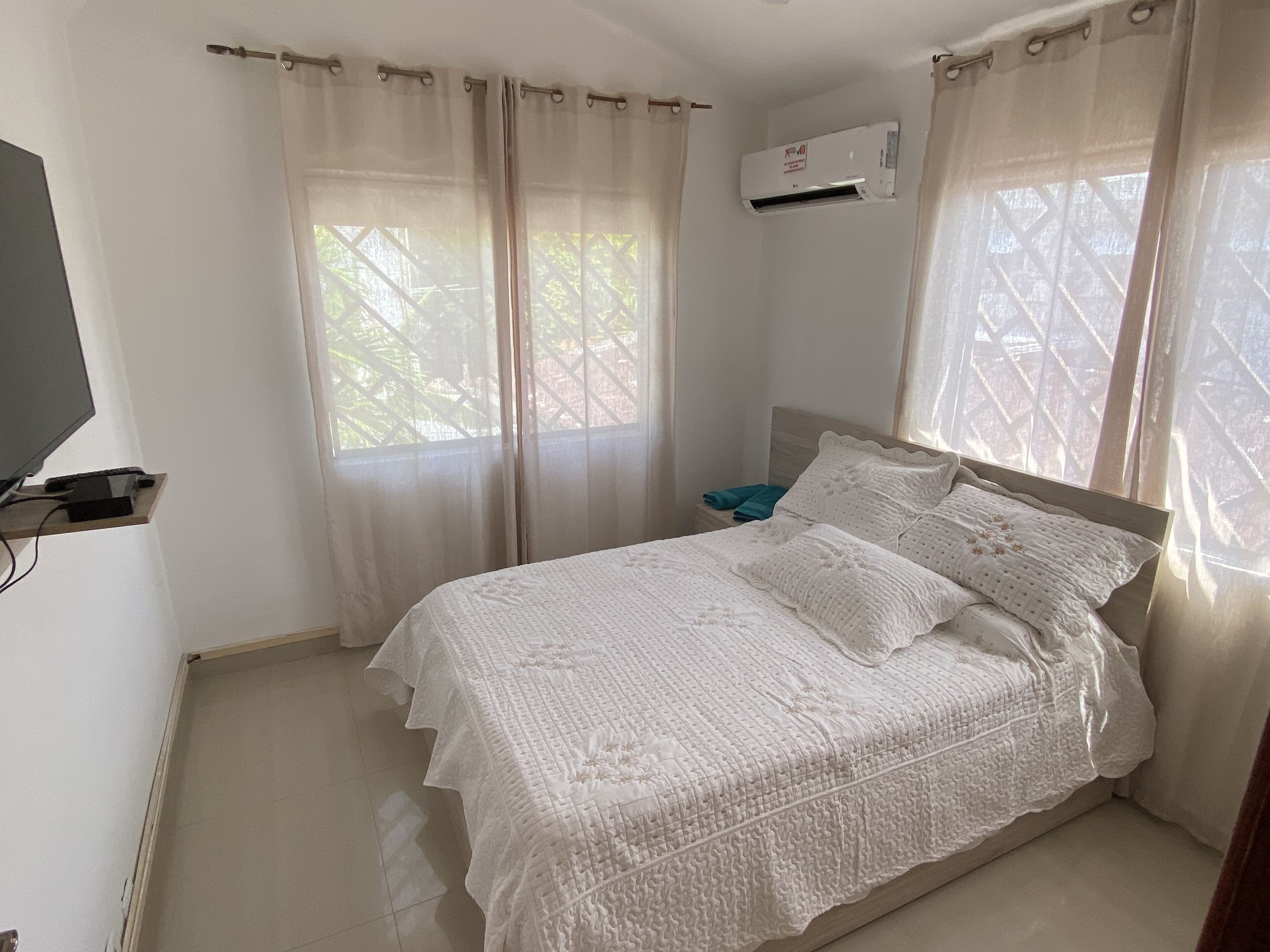 Un dormitorio con cama, cortinas, aire acondicionado, televisor y mesita de noche con una toalla.