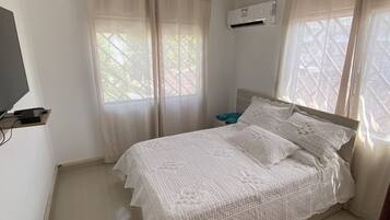 Un dormitorio con cama, cortinas, aire acondicionado, televisor y mesita de noche con una toalla.