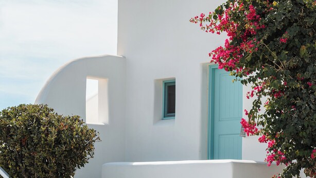 A white building with a blue door and a small window. There is a staircase leading up to the entrance. - Ploes Seaside Houses (Naxos)
