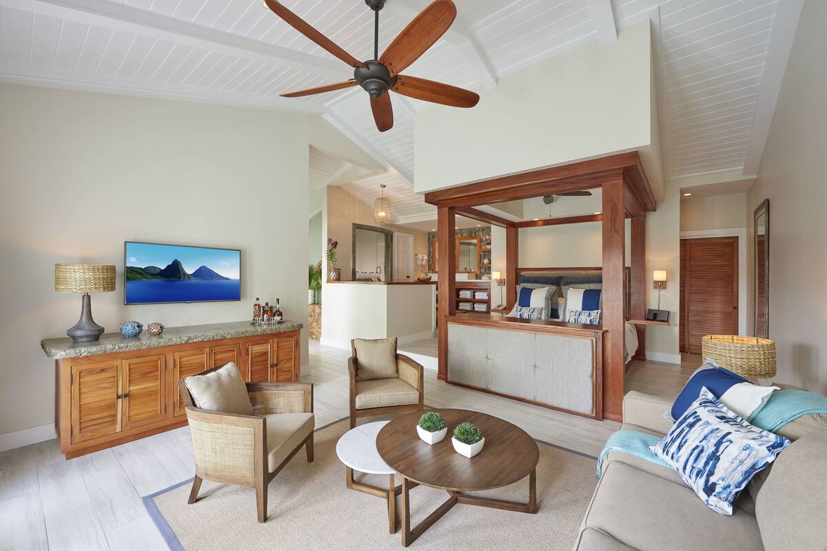 A spacious living room with a wooden ceiling fan, a large TV, and a kitchen area in the background.