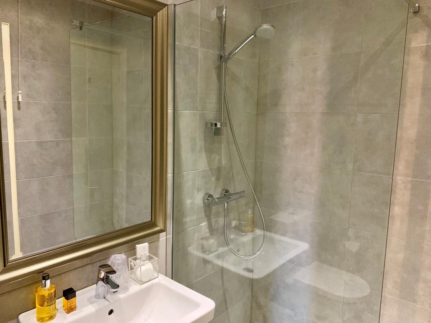 Moderni kylpyhuone, jossa on lasiseinällä varustettu suihku, valkoinen pesuallas ja hygieniatuotteet tasolla.
