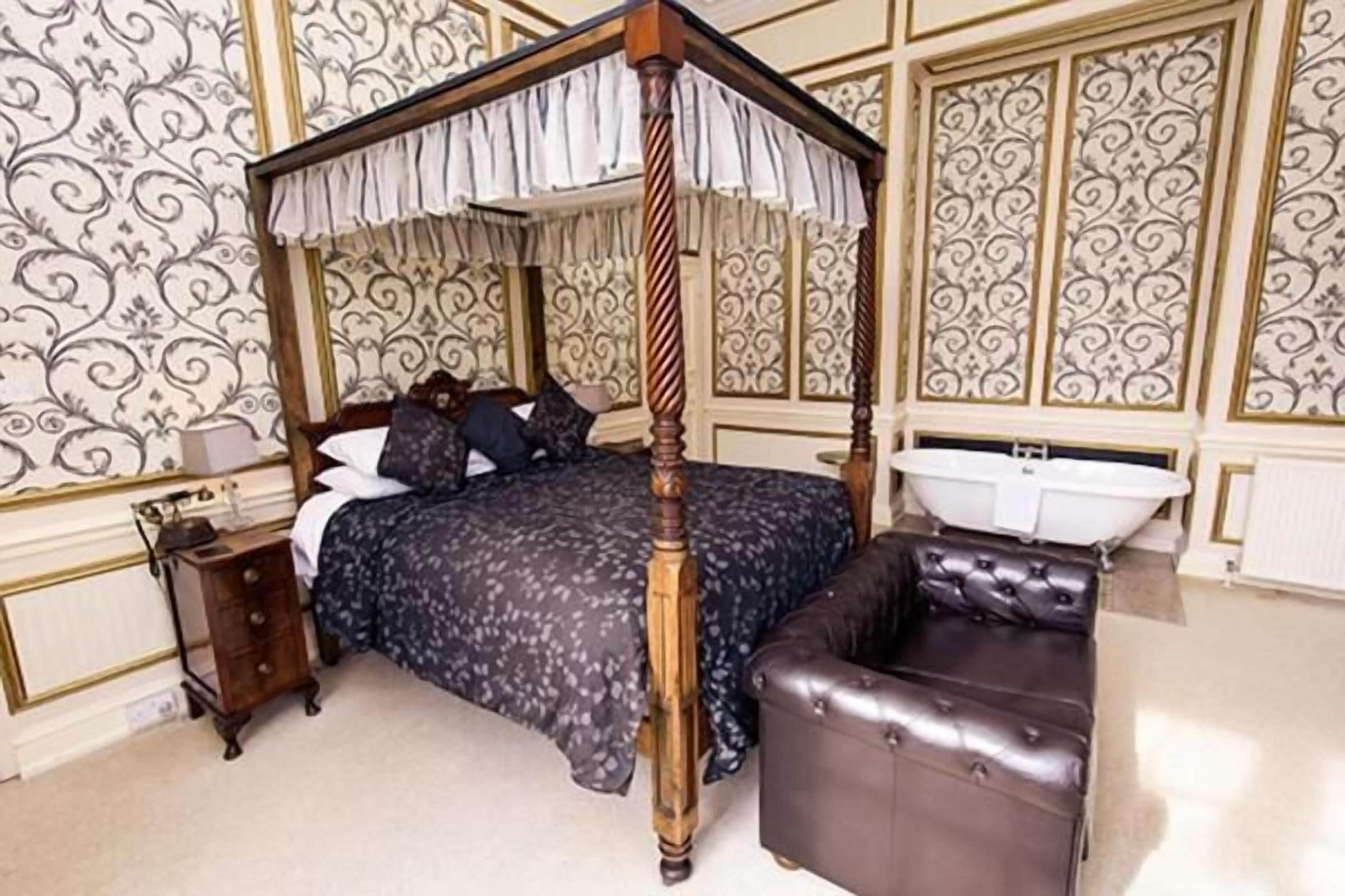 A four-poster canopy bed with a dark floral bedspread, a wooden nightstand, and a leather armchair.