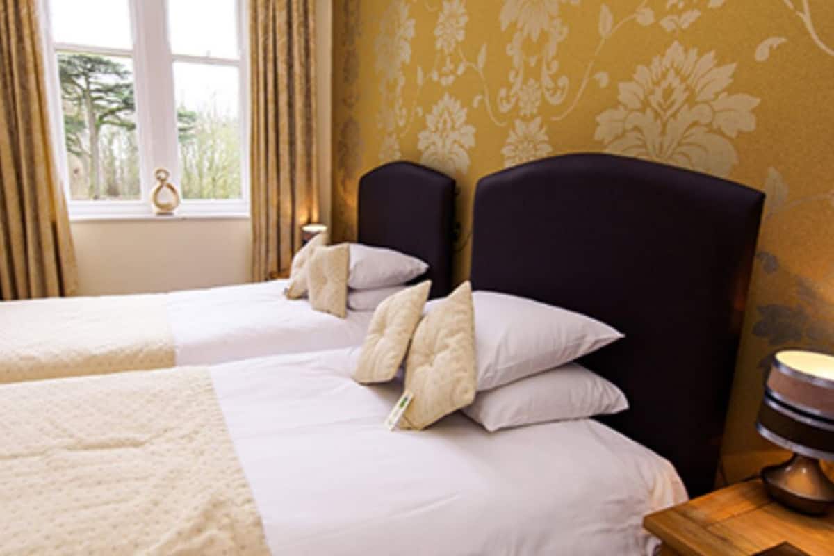 A hotel room with two beds, a window with curtains, a lamp on a bedside table, and a wall with a floral pattern.